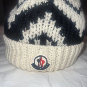 Stickad Moncler mössa med tofs - Snygg stickad mössa från Moncler i marinblått och vitt med coolt grafiskt mönster. Mössan har en stor vit tofs på toppen och uppvikt kant med Moncler-logga framtill. Perfekt för kalla dagar när du vill sticka ut lite extra.