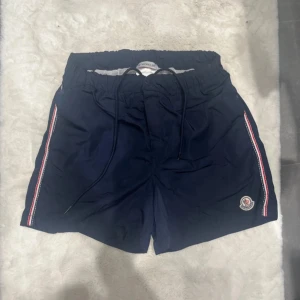 Marinblå Moncler shorts med snörning - Snygga marinblå shorts från Moncler med coola rödvita ränder längs sidorna och broderad logga. De har elastisk midja med snörning och meshfoder på insidan. Perfekta för sommaren och chill dagar vid vattnet.