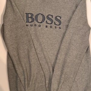 Grå tröja från Hugo Boss med logga - Snygg grå tröja från Hugo Boss med broderad BOSS-logga i svart och vitt framtill. Tröjan har rund halsringning, långa ärmar och är tillverkad i mjuk bomull. 