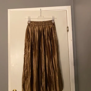 Plisserad guldbrun långkjol i satin - Säljer en plisserad långkjol i guldbrun satin med glansig finish. Kjolen har resår i midjan för skön passform och de djupa veck ger ett snyggt fall. Perfekt för dig som gillar att sticka ut med metallictoner och elegant vibe.⭐️