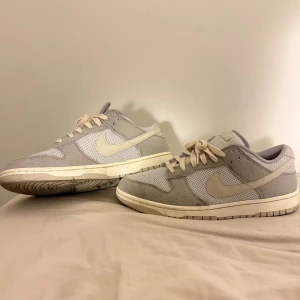 Nike Dunk Low i ljusgrå mesh och mocka - Nike Dunk Low sneakers i ljusgrå och beige färgton med meshpaneler och detaljer i mocka. Klassisk rund tå, platt sula och snörning. Swoosh-logga på sidan och låg siluett som passar till streetwear. Snygg och stilren design för dig som gillar neutrala färger.