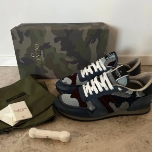 Valentino Garavani Rockrunner sneakers - Snygga Valentino Garavani Rockrunner sneakers med coolt camouflage-mönster i blått, grått och vinrött. Skorna har vit snörning, rund tå och platt sula. Materialmix av mocka och mesh ger en lyxig vibe. Perfekta för dig som vill sticka ut med designer-sneakers. 