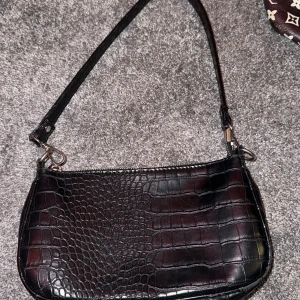 Svart axelväska i croco-imitation - Snygg svart axelväska från Gina Tricot med croco-imitation och stilren form. Väskan har ett huvudfack med dragkedja och en justerbar axelrem. Perfekt storlek för det viktigaste och ger en cool vibe till din outfit.