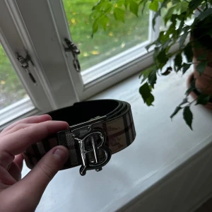 Rutig Burberry bälte  med silverdetalj - Snyggt Burberry-bälte med klassiskt rutigt mönster i beige, svart och rött. Bältet har en stor silvrig spänne med Burberrys ikoniska B-logga och är gjort i skinn. Perfekt accessoar för att lyfta vilken outfit som helst.