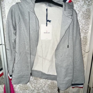 Grå Moncler hoodie med huva - Snygg grå hoodie från Moncler med dragkedja, huva och Moncler-logga på ärmen. Muddarna har stilrena ränder i blått, vitt och rött. Tillverkad i mjuk bomull, perfekt för chill dagar. Klassisk passform och riktigt clean look. Pris kan diskuteras 