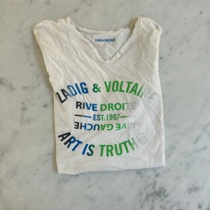 Vit t-shirt Zadig & Voltaire XS - Vit t-shirt från Zadig & Voltaire i storlek XS med tryck i blått, grönt och svart framtill. Modellen har korta ärmar, rund halsringning med två knappar och är gjord i mjuk bomull. Snygg och chill med coolt statement-tryck.