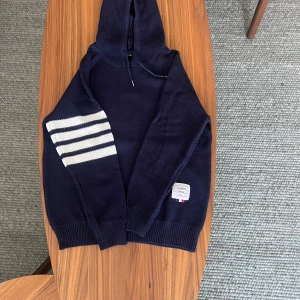 Mörkblå hoodie med vita ränder - Snygg mörkblå hoodie från Qingwai med fyra vita ränder på ena ärmen och patch med text framtill. Tröjan har huva med snörning och är stickad i ett grovt mönster. Perfekt för dig som gillar streetstyle och vill sticka ut lite extra.