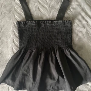 Svart smockad linnetopp - Säljer en svart linnetopp med breda axelband och smockad överdel. Toppen har en volangig nederdel 