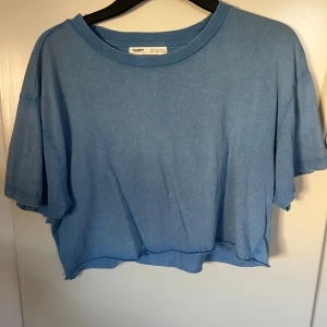 Blå croppad t-shirt från Pull&Bear - Säljer en blå croppad t-shirt från Pull&Bear i storlek M. T-shirten har en loose passform, korta ärmar och är tillverkad i mjuk bomull och har äcen washed mönster. Perfekt för en avslappnad och chill stil.