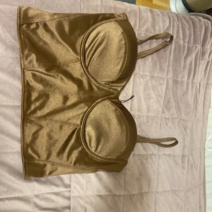 Bronsfärgad satin korsettopp - Säljer en snygg bronsfärgad korsettopp från NA-KD i glansigt satinmaterial. Toppen har smala axelband, formade kupor och dragkedja i ryggen för enkel på- och avtagning. Perfekt för dig som vill sticka ut med en trendig look.