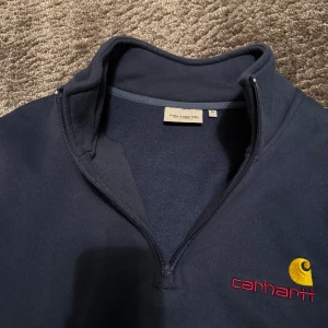 Mörkblå Carhartt quarter zip  - Säljer en mörkblå tröja från Carhartt med broderad logga i gult och rött på bröstet. Tröjan har half zip-dragkedja och hög krage, perfekt för lager-på-lager. Skön och stilren modell som funkar året runt.