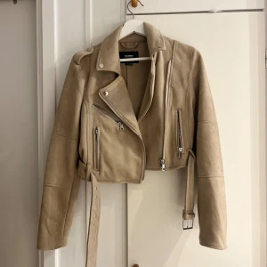 Beige mocka bikerjacka Pull&Bear - Snygg beige bikerjacka från Pull&Bear i mjuk mocka-look. Jackan har croppad passform, silvriga dragkedjor, axelklaffar och bälte i midjan. Perfekt för dig som vill ha en cool och stilren jacka med detaljer som sticker ut.