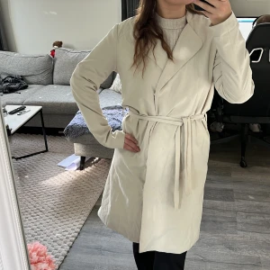 Beige trenchcoat från Noisy May  - Stilren beige trenchcoat från noisy may med klassisk krage och knäppning framtill. Lång modell med raka ärmar och lätt glansigt material som ger en snygg look. 