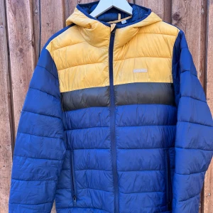 Blå och gul pufferjacka Jack & Jones - Fet pufferjacka från Jack & Jones i blått och gult med svarta detaljer. Jackan har huva, dragkedja framtill och två sidofickor med dragkedja. Perfekt för kalla dagar när du vill sticka ut lite extra.