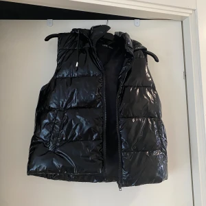 Svart glansig dunväst från SHEIN - Svart dunväst från SHEIN i storlek S med glansig finish och hög krage. Västen har dragkedja framtill och två fickor på sidorna. Perfekt för lager-på-lager och ger en cool vibe till din outfit. Aldrig använd! 