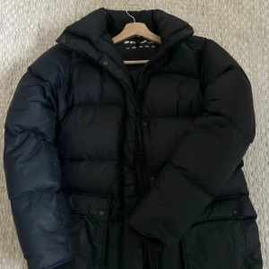 Svart pufferjacka från Everest - Svart pufferjacka från Everest med hög krage och dragkedja framtill. Jackan har stora fickor med knappar och snörning i midjan för en snygg passform. 80%dun.