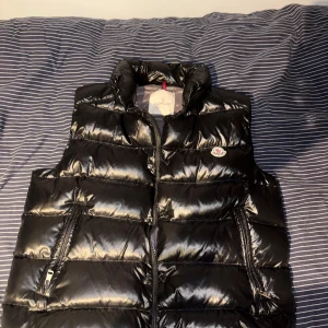 Svart dunväst från Moncler - Svart, glansig dunväst från Moncler med quiltad design och hög krage. Västen har två fickor med dragkedja och Moncler-logga på bröstet. Insidan har ett coolt serietidningsmönster och etikett med certifiering. Perfekt för kalla dagar.