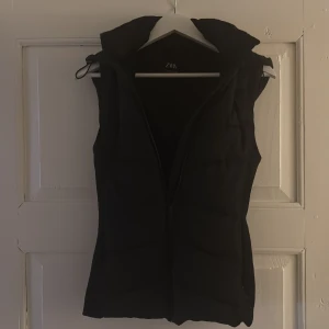 Svart dunväst med huva från Zara - Svart dunväst från Zara med snygg quiltad design och praktisk huva. Västen har dragkedja framtill och sidofickor med dragkedja. Perfekt för lager-på-lager och ger en cool, sportig vibe. Passformen är normal och materialet är polyester.