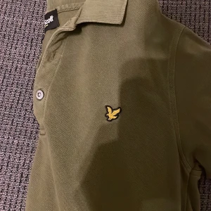 Olivgrön pikétröja från Lyle & Scott - Säljer en olivgrön pikétröja från Lyle & Scott med klassisk krage och två knappar framtill. Tröjan har det ikoniska gula fågelmärket broderat på bröstet och är gjord i mjuk bomull. Perfekt för dig som gillar stilren och enkel design.