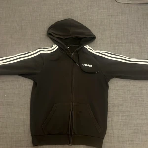 Svart Adidas hoodie med vita ränder - Svart hoodie från Adidas med klassiska vita ränder längs ärmarna och Adidas-logga på bröstet. Tröjan har huva med snörning, dragkedja framtill och två fickor. Perfekt för dig som gillar sportig och chill stil.