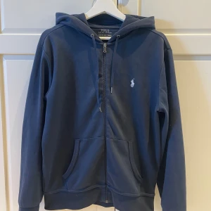 Mörkblå hoodie från Polo Ralph Lauren - Snygg mörkblå hoodie från Polo Ralph Lauren med dragkedja och huva. Klassisk logga broderad på bröstet och praktiska fickor framtill. Tillverkad i mjuk bomull, perfekt för chill dagar. Långärmad och med snörning i huvan.