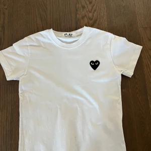 Vit t-shirt Comme des Garcon - Säljer en clean vit t-shirt från Comme des Garcons Play i storlek 150 cm med det ikoniska svarta hjärtat med ögon broderat på bröstet. Klassisk rund hals och korta ärmar.