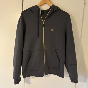 Mörkgrå hoodie med dragkedja från BOSS - Säljer en stilren mörkgrå hoodie från BOSS med guldfärgad dragkedja och logga på bröstet. Tröjan har huva med snörning, två stora fickor framtill och ribbade muddar. Perfekt för en chill och snygg look. Har en liten fläck på baksidan