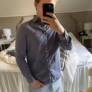 Blå skjorta från Ralph Lauren - Snygg blå skjorta från Ralph Lauren med klassisk krage och knappar hela vägen. Den har en liten röd broderad logga på bröstet och långa ärmar med knapp vid manschetten. Perfekt för dig som gillar stilrena och tidlösa plagg.