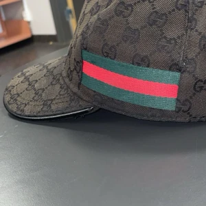 Gucci keps med GG-mönster och röd/grön rand - Snygg keps från Gucci i mörkbrunt tyg med klassiskt GG-monogram över hela kepsen. På sidan finns den ikoniska gröna och röda randen. Böjd skärm och justerbar passform. Perfekt accessoar för dig som vill sticka ut med lyxig streetstyle.