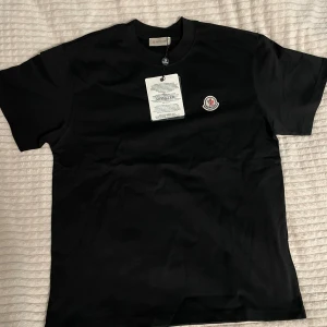 Moncler svart och vit t-shirt - Snygga t-shirts från Moncler i svart och vitt med klassisk rund hals och korta ärmar. På bröstet sitter Monclers ikoniska logotyp-patch. Materialet är mjuk bomull som känns skönt mot huden. Perfekt för dig som gillar stilrena plagg