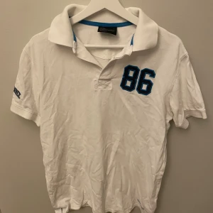 Vit pikétröja Peak Performance - Vit pikétröja från Peak Performance med blå detaljer och broderad siffra 86 på bröstet. Klassisk krage, kortärmad och med broderad logga på ärmen. Tillverkad i bomull och perfekt för en sportig och clean stil.