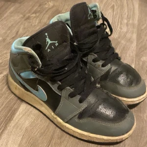 Nike Air Jordan 1 High svart/grå - Nike Air Jordan 1 High sneakers i svart och grått med ljusblå detaljer. Klassisk high-top siluett med snörning och rund tå. Skorna har en slitstark gummisula och är tillverkade i skinn. Ikonisk Jumpman-logga på plösen och Swoosh på sidan.