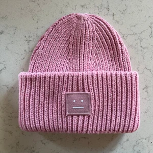 Rosa stickad mössa från Acne Studios - Säljer en rosa ribbstickad mössa från Acne Studios med det ikoniska ansikts-emblemet framtill. Mössan är tillverkad i mjukt ullmaterial och har bred uppvikt kant. Perfekt för dig som vill ha en cool och trendig accessoar. Hör av dig vid minsta fråga!