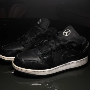 Nike Air Jordan 1 Low Svart/Vit 40 - Säljer ett par Nike Air Jordan 1 Low i svart med vita detaljer och broderad Air Jordan-logga på hälen. Skorna har klassisk snörning, rund tå och vit mellansula. Tillverkade i skinn och textil för en clean och tidlös look. Perfekta för dig som gillar streetwear och sneakers.