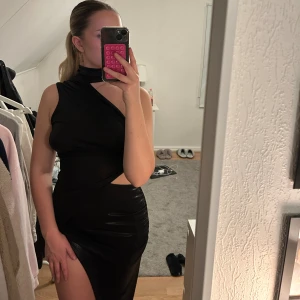 Svart oneshoulder klänning med slits - Säljer en svart, glansig oneshoulder klänning med hög slits och cut-out vid midjan. Klänningen har en tight passform och en snygg halsdetalj. Använd endast vid ett tillfälle.