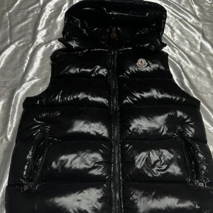 Svart dunväst från Moncler - Snygg svart dunväst från Moncler med glansig finish och klassisk logga på bröstet. Västen har huva, två dragkedjefickor och hel dragkedja framtill. Perfekt för lager-på-lager och ger en clean streetstyle-vibe.