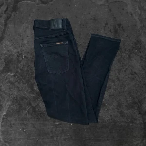 Svarta jeans från Nudie Jeans  - Svarta jeans från Nudie Jeans i klassisk femficksmodell. Jeansen har en smal passform med raka ben och är tillverkade i ett mjukt bomullstyg med stretch. Snygga detaljer med svarta sömmar och diskret logga på bakfickan. Perfekta för en clean och stilren look.