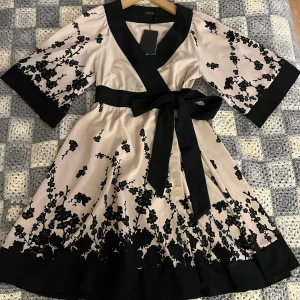 Blommig kortklänning från Stockh LM - Superfin kortklänning från Stockh LM i ljus champagnefärg med svarta blommiga broderier. Klänningen har trekvartsärm, knyts omlott med bred svart kant och knytband i midjan. Tillverkad i glansigt material som ger en lyxig känsla. Inköpt i sommar för 599, aldrig använd.