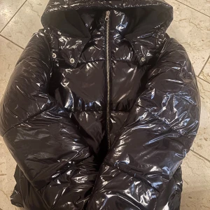 Svart glansig pufferjacka från Nelly - Svart pufferjacka från Nelly med glansig finish och huva. Jackan har dragkedja framtill, stora fickor och quiltad design. Perfekt för kalla dagar! Använd minst 2 ggr. Inga tecken på användning💕