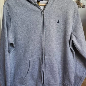 Grå zip hoodie från Polo Ralph Lauren i ungdoms XL - Klassisk grå zip hoodie från Polo Ralph Lauren i storlek ungdoms XL/170. Tröjan har dragkedja framtill, huva och kängurufickor. Liten mörkblå logga broderad på bröstet. Perfekt för chill dagar och enkel att matcha med jeans eller joggers.