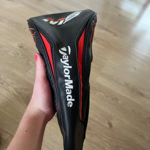 TaylorMade M6 Headcover - TaylorMade M6 headcover i svart och rött, passar fairway woods eller hybrider. Snygg design med tydlig logotyp och modellbeteckning. I mycket fint skick utan synligt slitage eller skador. Perfekt för att skydda din klubba och ge din golfbag ett proffsigt utseende.