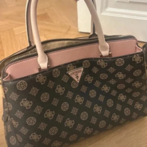 Svart handväska från Guess med mönster - Snygg svart handväska från Guess med beige och rosa detaljer. Väskan har ett unikt mönster med loggor och symboler, ljusrosa handtag och insida. Praktisk dragkedja upptill och flera fack för dina grejer. Perfekt för dig som gillar stil och funktion.