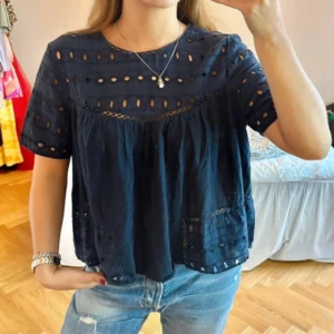 Mörkblå blus med broderade detaljer - Säljer en mörkblå blus med korta ärmar och broderade håldetaljer. Blusen har en lös passform och rund halsringning. Perfekt att matcha med jeans för en avslappnad look.