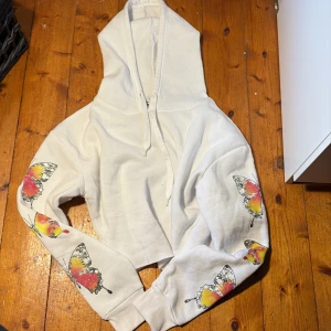 Vit hoodie med fjärilar från Roma Concept - Vit croppad hoodie från Roma Concept med stora färgglada fjärilar i rött, gult och svart tryck på både ärmar och rygg. Tröjan har huva med snörning och ribbade muddar. 