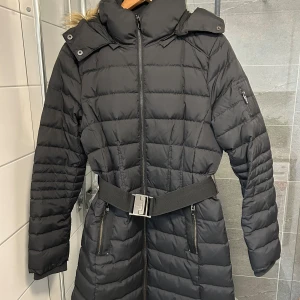 Svart lång dunjacka från Esprit - Säljer en svart lång dunjacka från Esprit med huva och snygg fuskpäls. Jackan har quiltad design, dragkedja framtill, två fickor med dragkedja och ett brett bälte i midjan. Perfekt för kalla dagar och har ribbade muddar vid ärmslut.