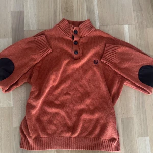 Orange stickad tröja från Chaps - Säljer en orange stickad tröja från Chaps med ribbad krage och knappslå med fyra knappar. Tröjan har förstärkta armbågar i mörkt tyg och broderad logga på bröstet. Perfekt för dig som gillar klassisk stil med en twist.