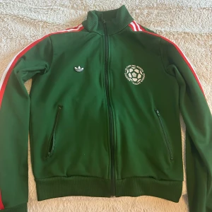 Grön Adidas track jacket Mexico 70 - Retroinspirerad grön Adidas track jacket med vita och röda ränder längs ärmarna, broderad logga och tryck med 'Mexico 70' på ryggen. Väldigt skön tröja. Storlek xs-s. 