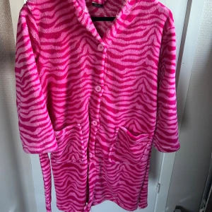 Sötaste zebramorgonrocken! - Mysig rosa fleecemorgonrock från KappAhl, med zebramönster och roliga detaljer som huva med broderat djuransikte och öron. Den har knappar framtill, två stora fickor och fastsytt bälte i midjan. 