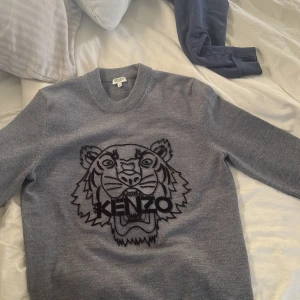 Grå stickad Kenzo tröja med tiger - Säljer en grå stickad tröja från Kenzo med ikoniskt svart broderat tigermotiv och logga på bröstet. Tröjan har rund halsringning och långa ärmar, perfekt för dig som vill ha en stilren men ändå statement look.