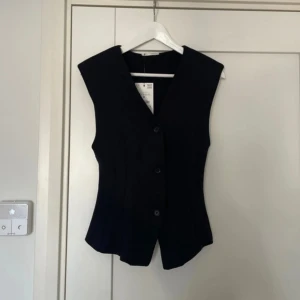 Svart figursydd väst från Zara - Snygg svart väst från Zara i figursydd modell med v-ringning och tre knappar framtill. Västen har stilrena sömmar som ger en cool siluett och är tillverkad i ett slätt polyestermaterial. Perfekt att styla över en t-shirt eller skjorta för en trendig look.